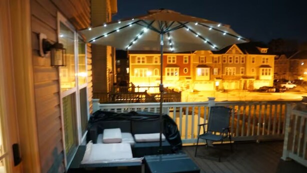 Une terrasse en bois avec des sièges extérieurs, un espace couvert illuminé de guirlandes lumineuses et un bâtiment en arrière-plan.