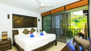 A bedroom with a large bed, a ceiling fan, a nightstand, and a view of greenery through the window.