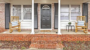 A front porch with a black door, two rocking chairs, a small table with a plant, and a welcome mat. - Quiet Comfort  Chas Attractions- Family Friendly (Goose Creek)