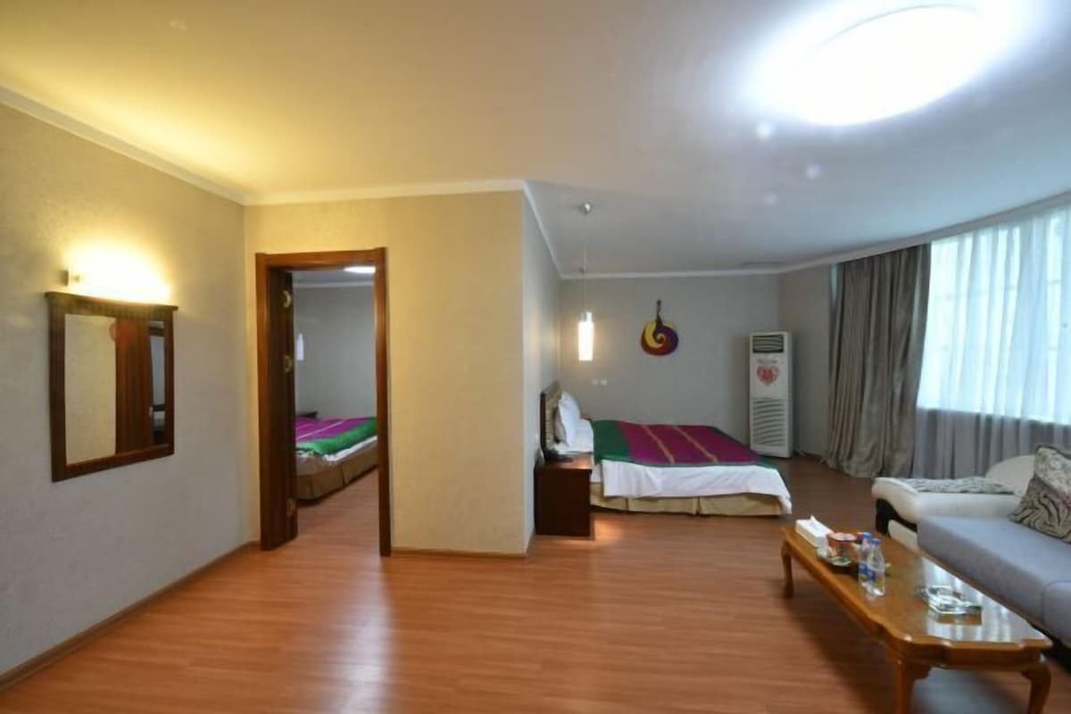 A hotel room with a wooden floor, a bed with a colorful blanket, a sofa, a small table with a bottle, a mirror, and a wall-mounted heater.