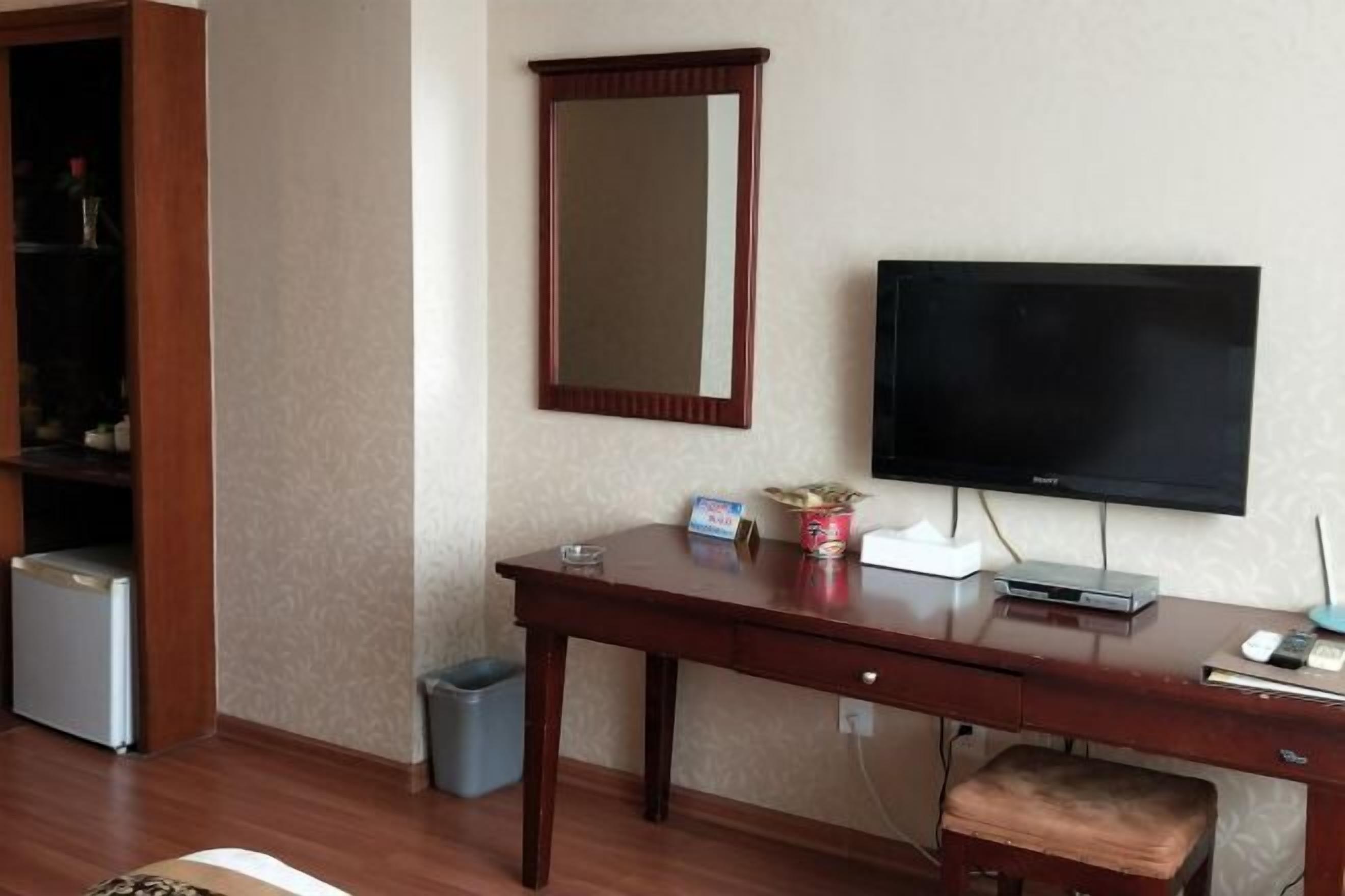 A hotel room with a wooden desk, a television, a small trash bin, and a cabinet with a mirror.