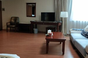 A hotel room with a wooden floor, a desk with a television, a chair, a sofa, and a lamp.
