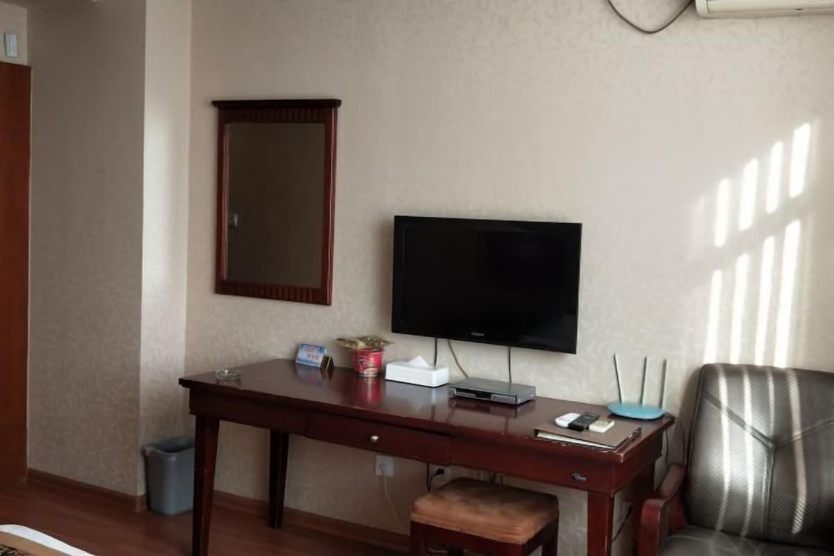 A hotel room with a wooden desk, a flat-screen TV, a mirror, a trash bin, and a leather chair.