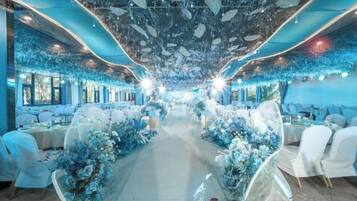 Une salle de banquet où les tables sont disposées de part et d’autre d’une allée centrale, entourées de compositions florales et de chaises blanches.