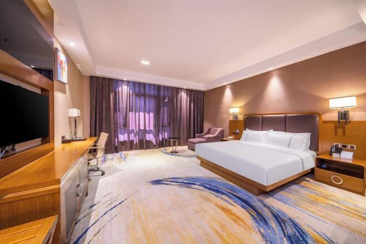 Quarto de hotel com uma cama grande, uma escrivaninha com cadeira, televisão e uma janela ampla com cortinas.