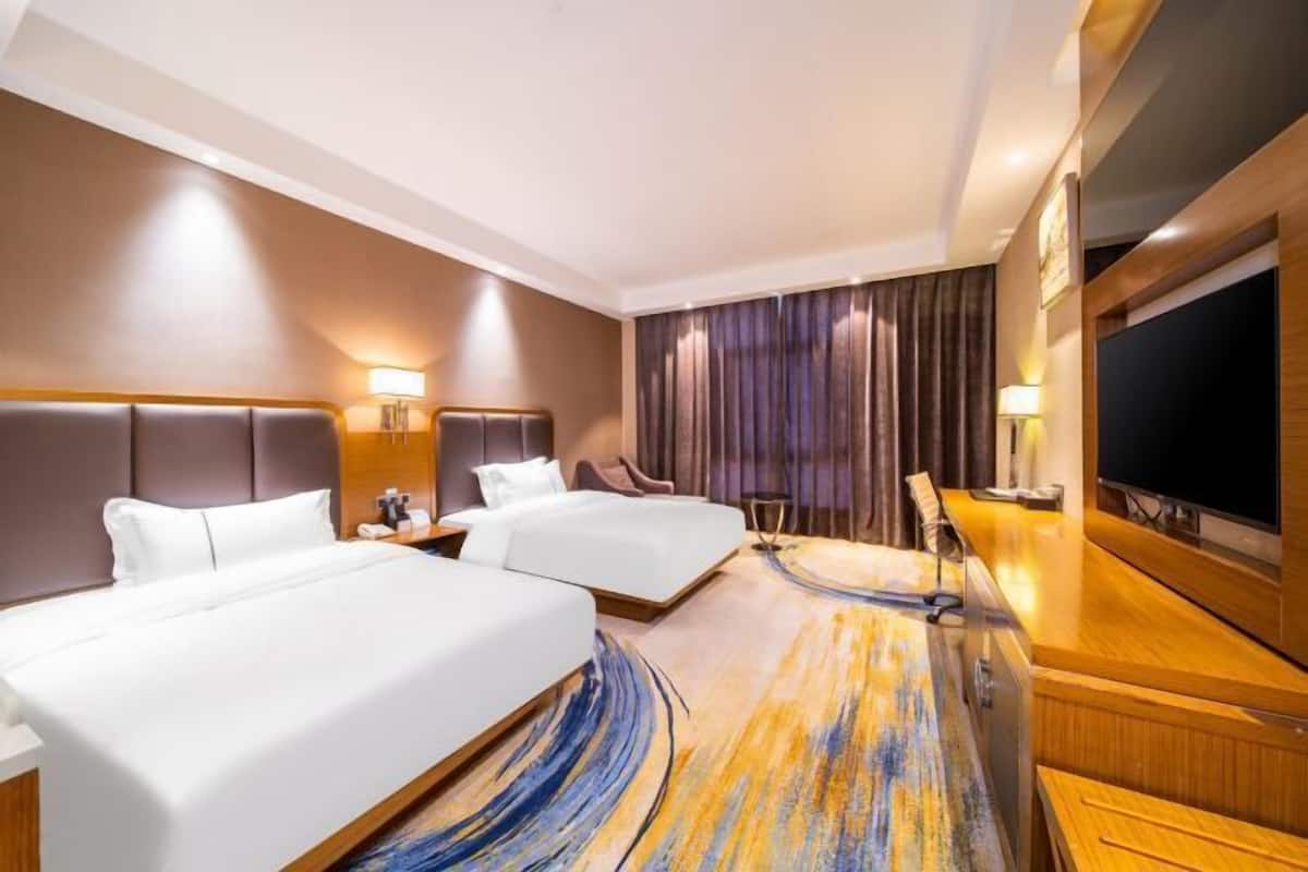Quarto de hotel com duas camas, televisão, escrivaninha e uma grande janela com cortinas.