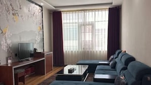 Un salon moderne avec un long canapé bleu foncé, un téléviseur à écran plat posé sur un meuble en bois et une grande fenêtre ornée de rideaux légers.