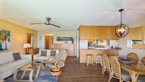 A spacious living area with a sofa, a glass coffee table, wicker chairs, a dining table with chairs, a kitchen area, and a ceiling fan. - Maui Sunset Vacation Rentals A419 (Kihei)