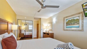 A hotel room with a bed, a ceiling fan, a wooden nightstand, a lamp, a mirror, and a framed picture on the wall. - Maui Sunset Vacation Rentals A419 (Kihei)