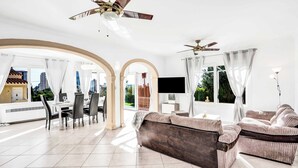 Uma sala de estar ampla com sofá, mesa de jantar e cadeiras, janelas grandes com cortinas e ventiladores de teto.