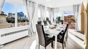 Uma sala de jantar com mesa posta para seis pessoas, grandes janelas com cortinas e vista para uma paisagem urbana e montanhas.