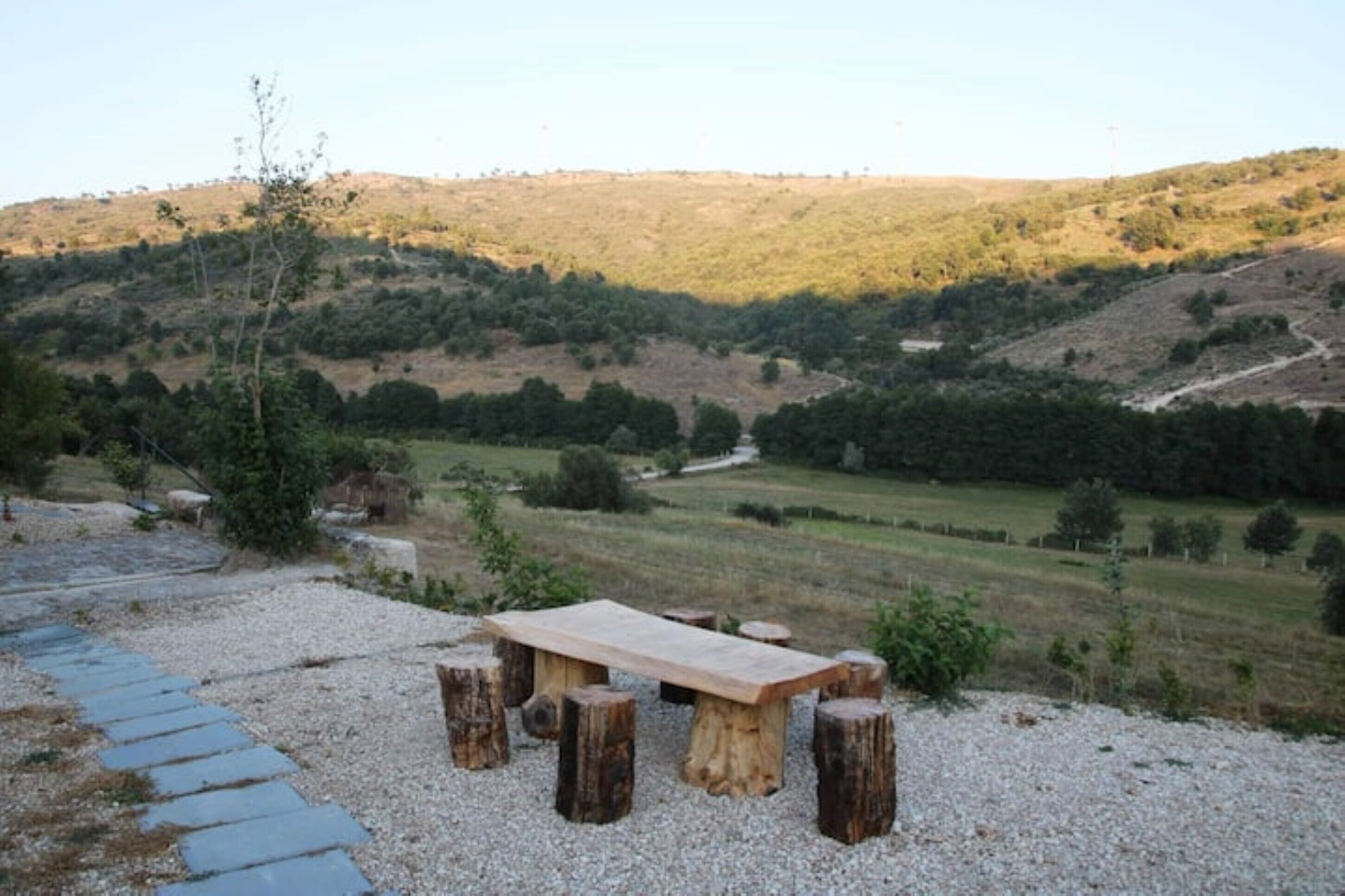 A rustic outdoor seating area with a wooden table and benches, surrounded by gravel and a scenic hillside view.