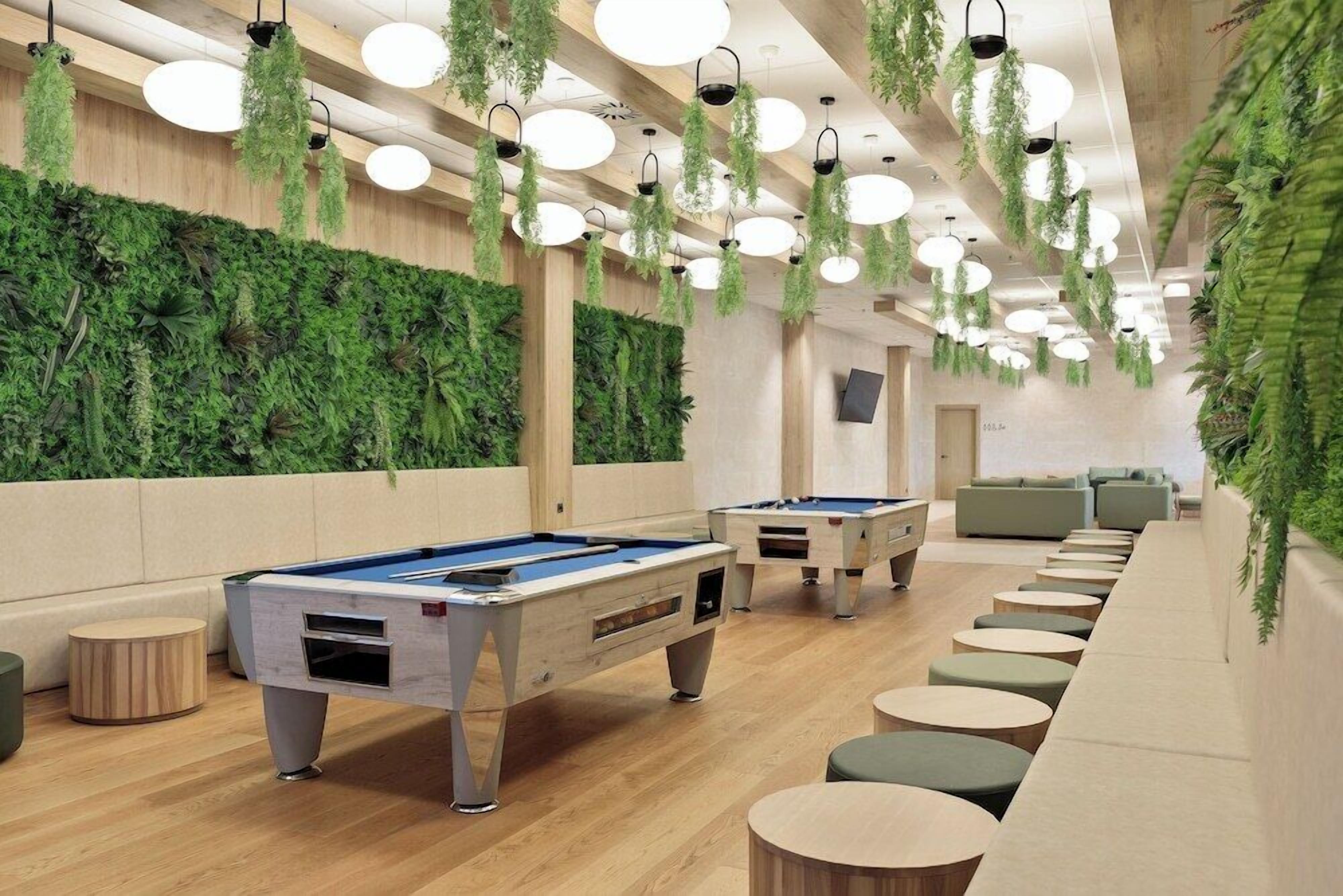 Una sala de billar con varias mesas de billar, plantas colgantes y un área de estar con mesas redondas.