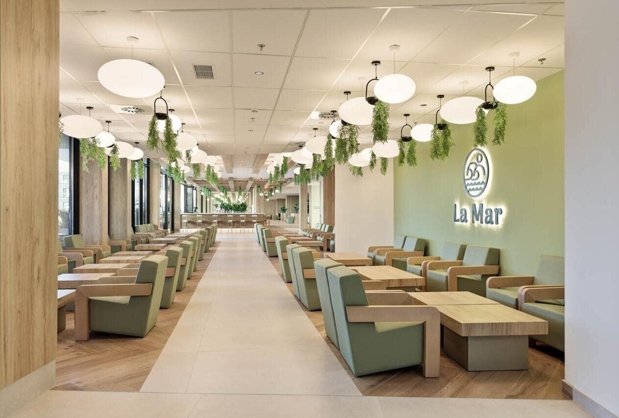 A modern restaurant interior with long tables and chairs, hanging plants, and a green wall with the restaurant's logo.