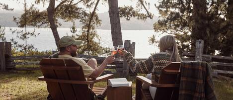 Two people sitting on wooden chairs outdoors with a view of a body of water and trees.