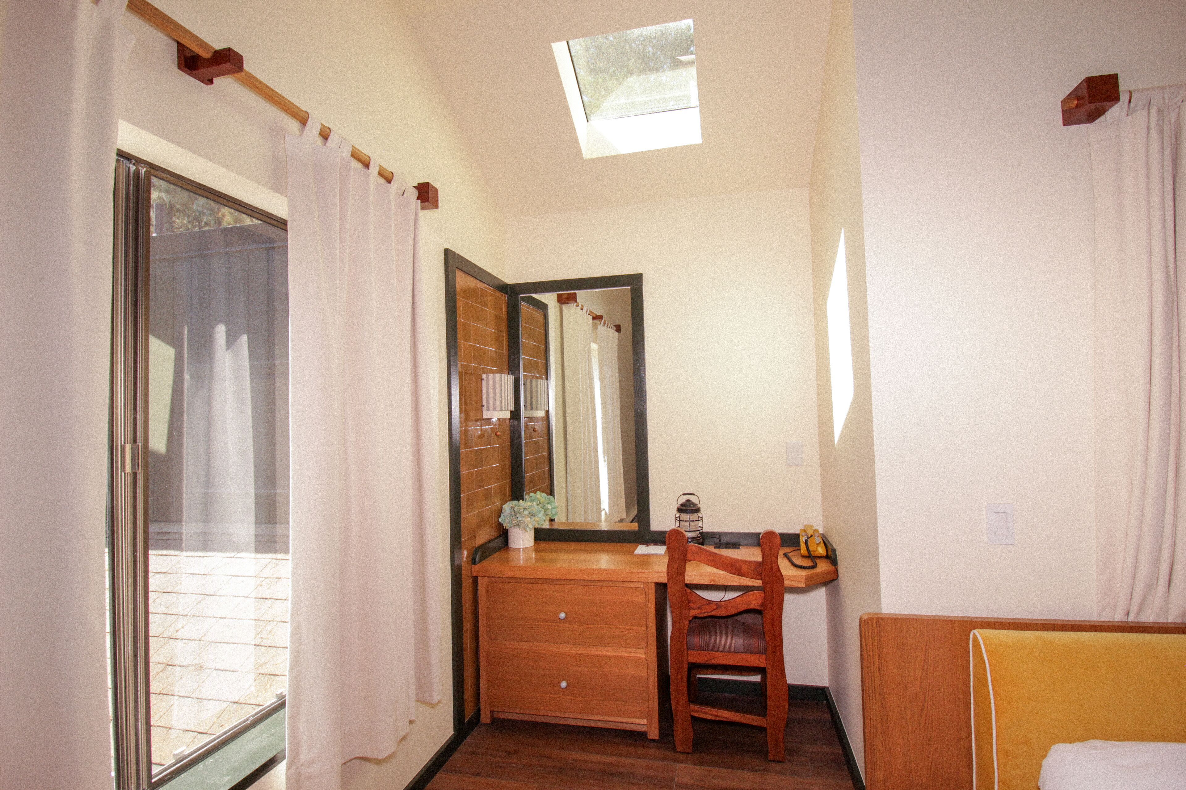 A room with a wooden desk, a chair, a mirror, a window with curtains, and a skylight.