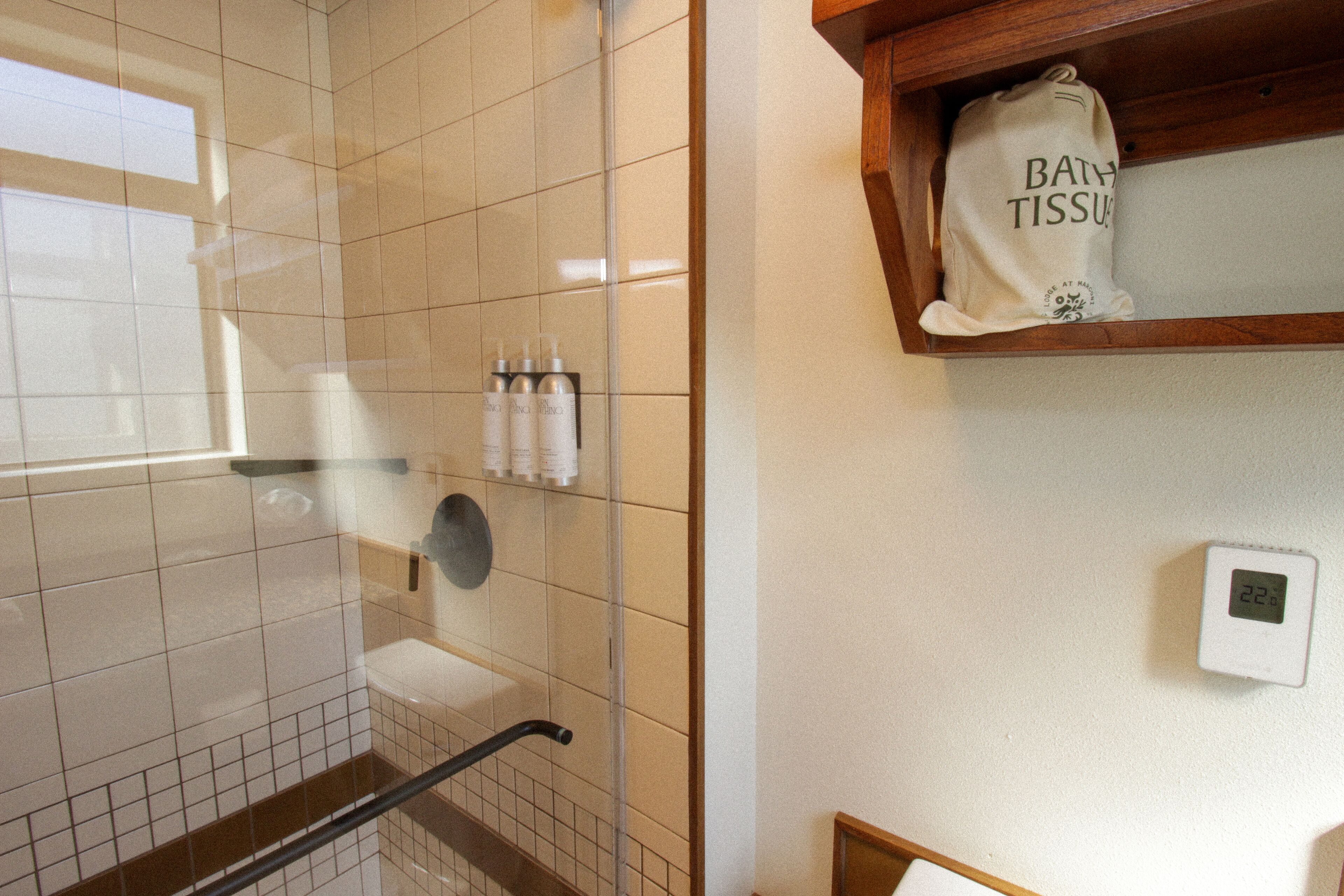 A bathroom with a glass shower enclosure, a wooden shelf with a bath tissue package, and a wall-mounted thermostat.