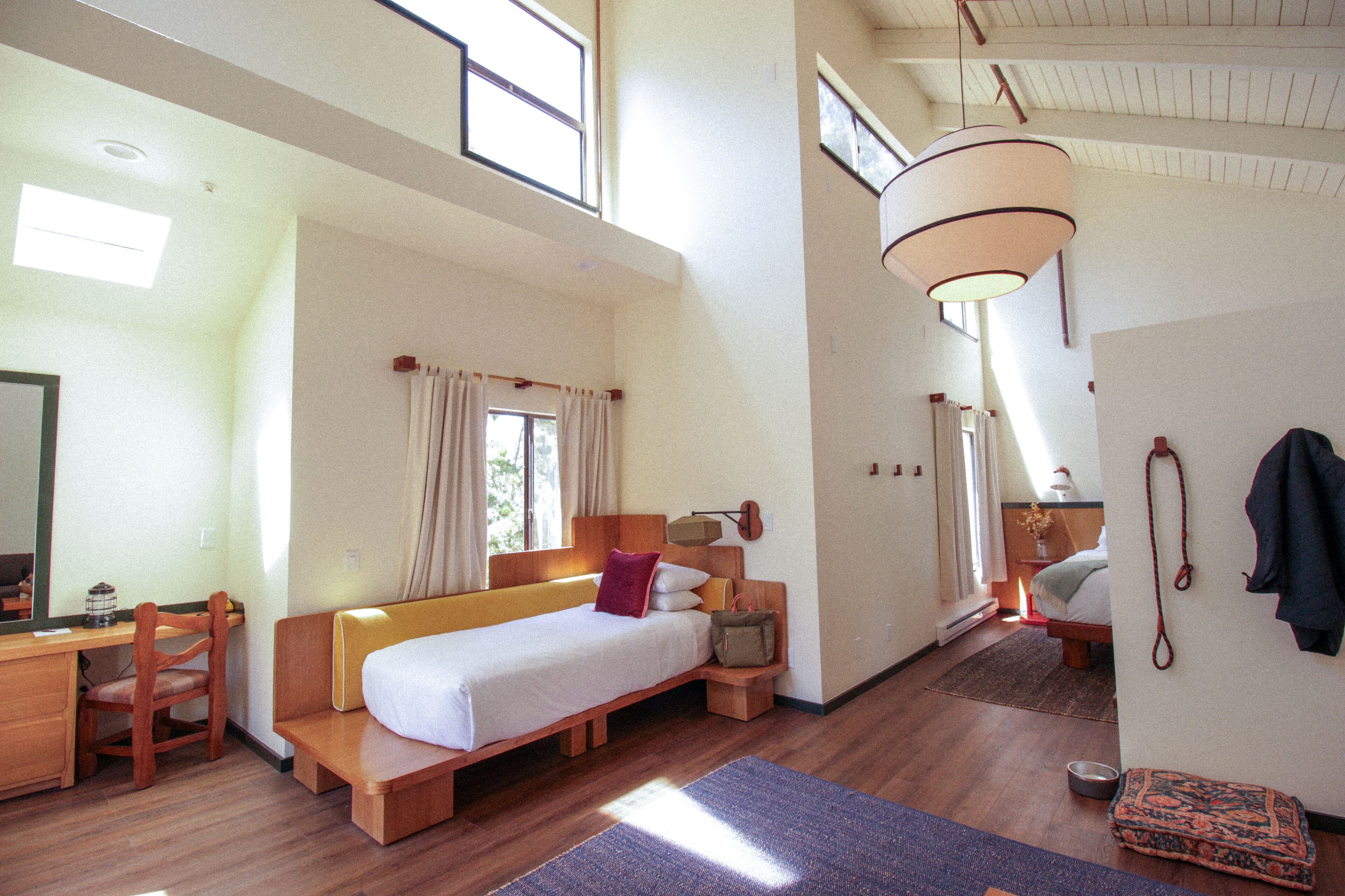 A spacious room with a large bed, a desk, and a chair. There is a window providing natural light, and a hanging light fixture.