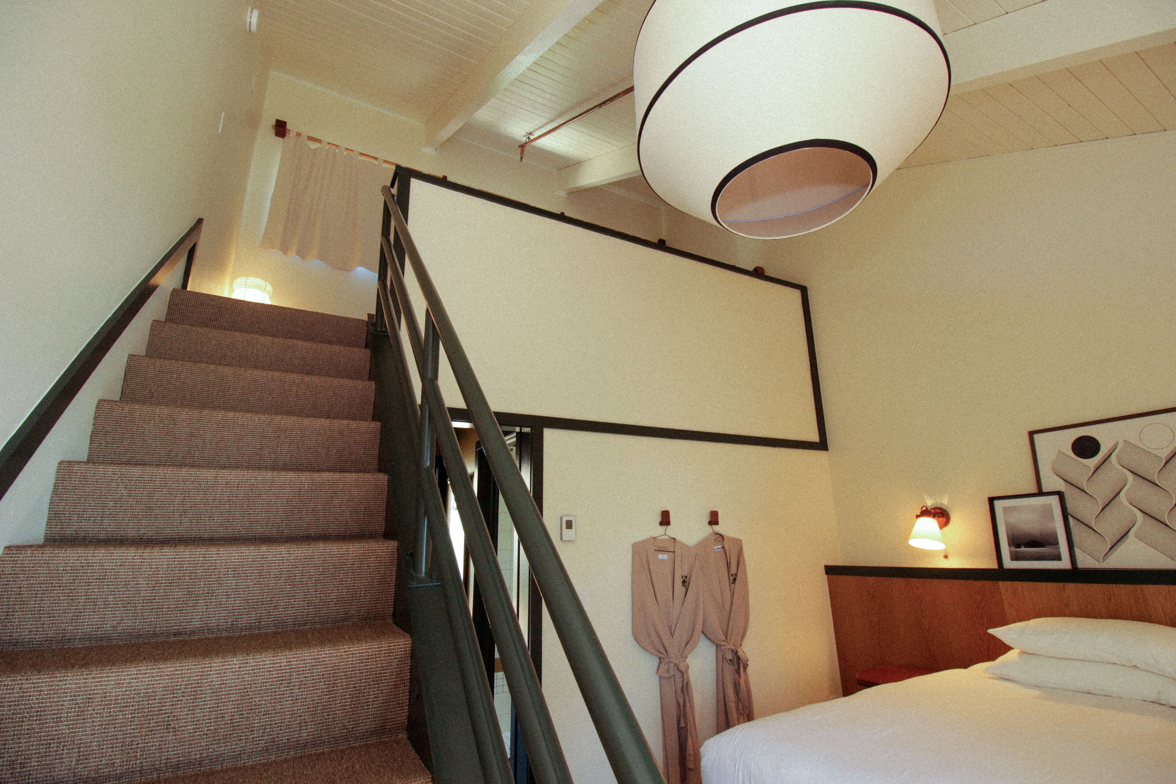 Habitación de hotel con una escalera, una cama, una mesita de noche, una lámpara y una pared decorada con un cuadro y un objeto enmarcado.