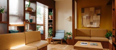 Ein gemütliches Wohnzimmer mit Holzmöbeln, einer Couch, einem Sessel und einem Couchtisch. Übertöpfe mit Pflanzen und ein Wandregal mit Büchern und Dekorationsobjekten.