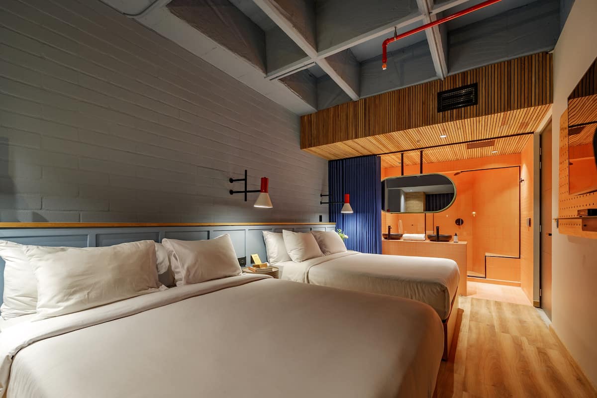 A modern hotel room with two beds, a wooden ceiling, and a bathroom with a mirror and orange tiles.