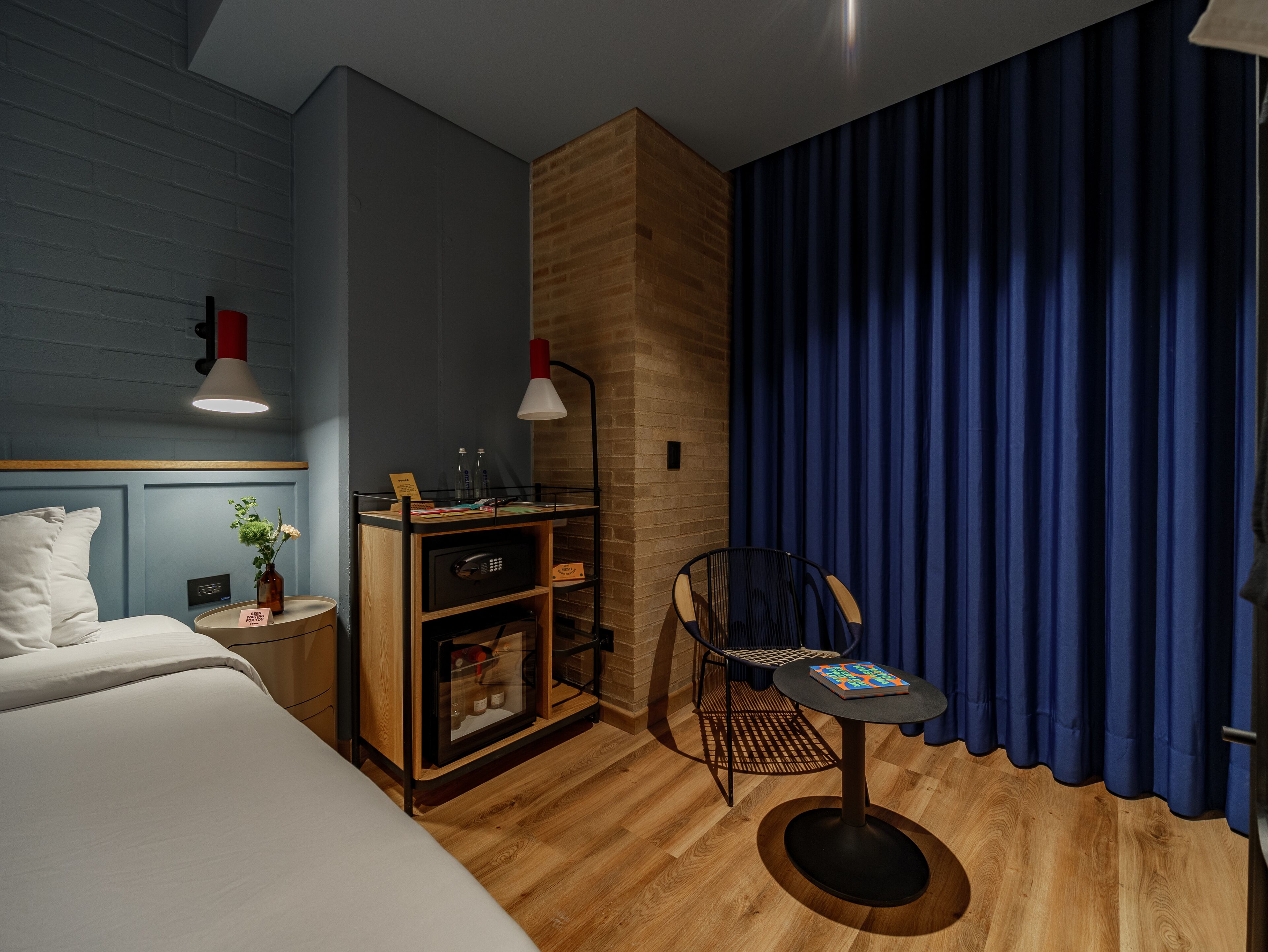 A hotel room with a bed, a desk with a lamp, a chair, and a small table with a book.