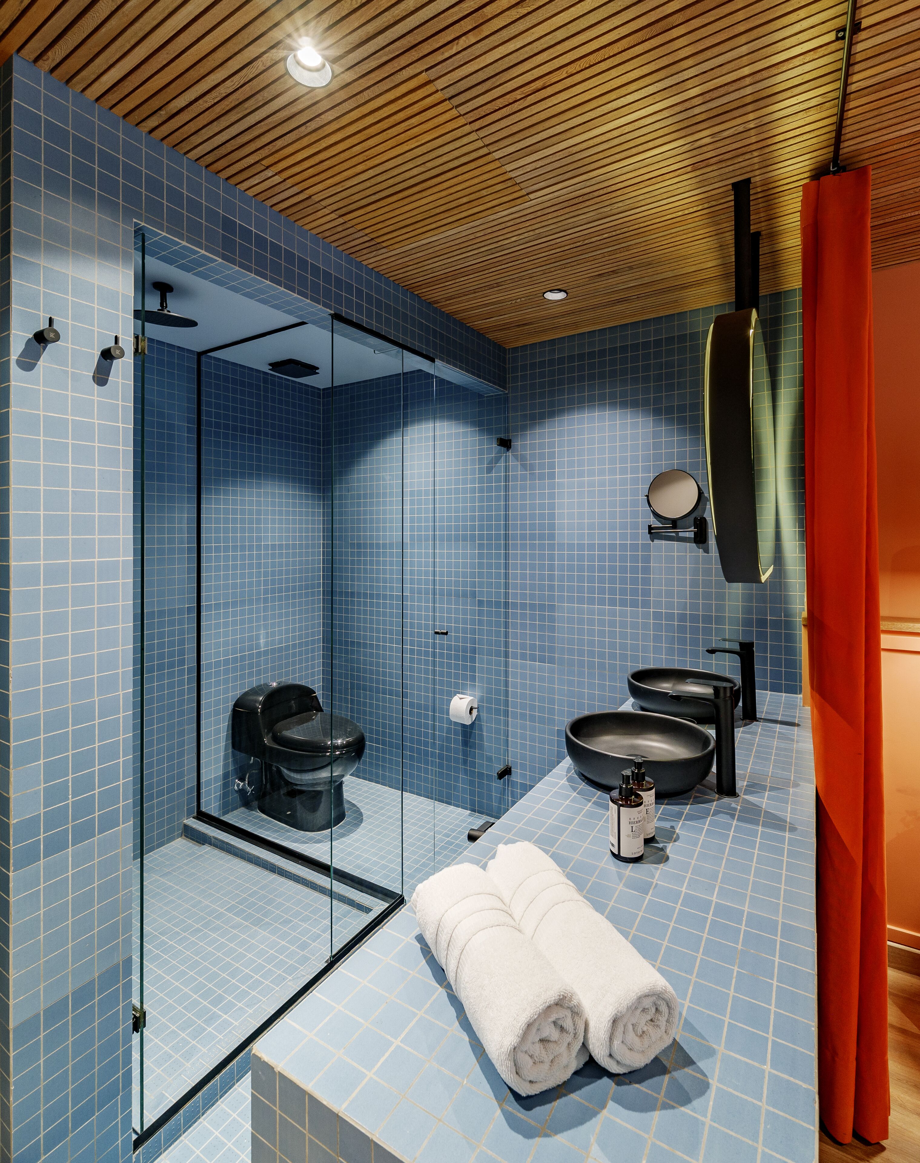 Un baño moderno con azulejos azules, una cabina de ducha de vidrio, un inodoro negro, un lavabo negro grande y una toalla en el suelo.