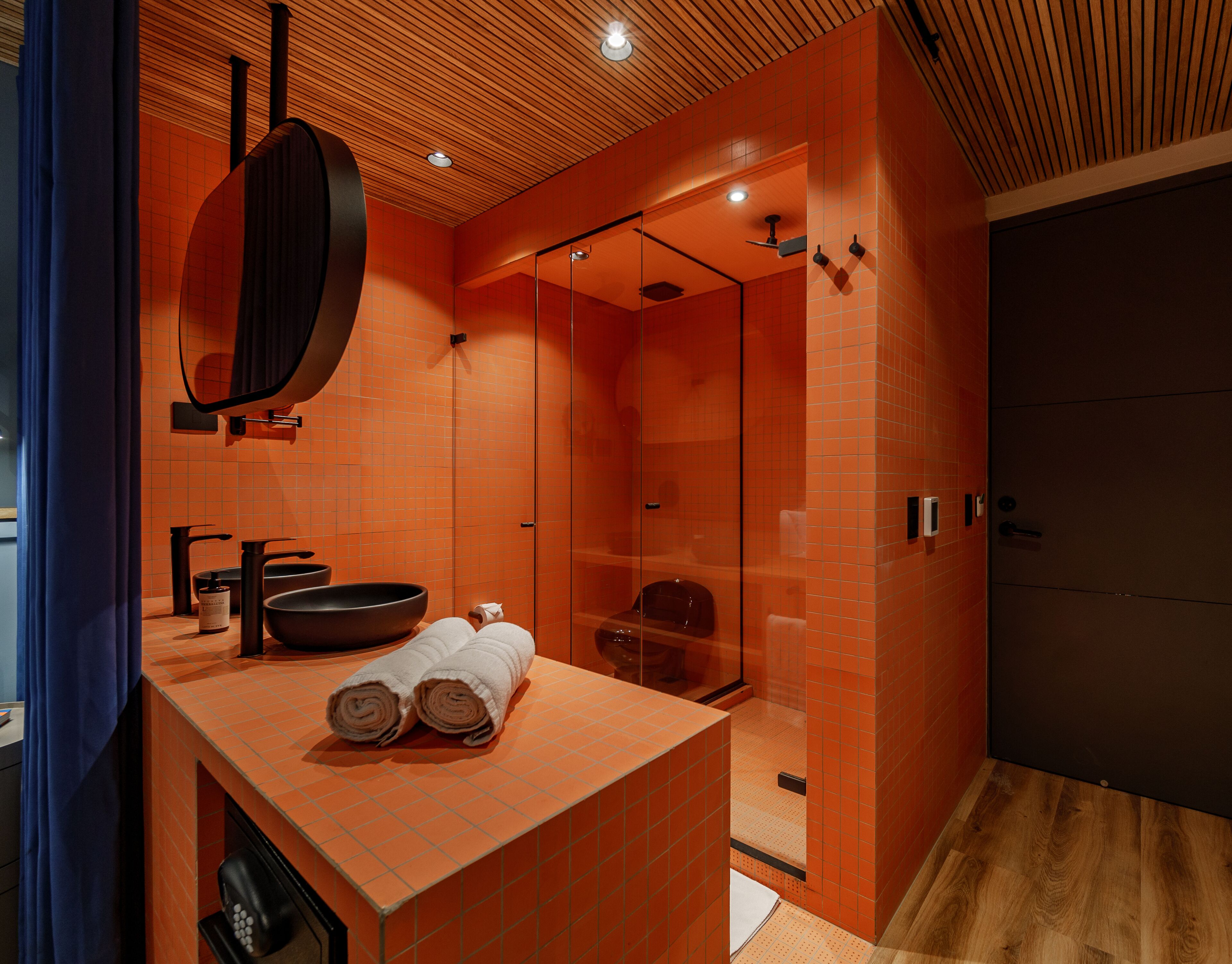 A modern bathroom with orange tiles, a wooden ceiling, a large mirror, a black sink, and a glass shower enclosure.
