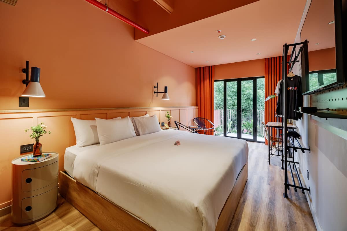 A modern hotel room with a large bed, a desk with a chair, a TV, and a balcony with orange curtains.