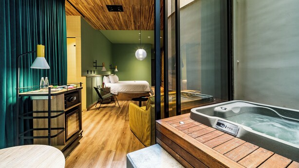 Una habitación de hotel moderna con techo de madera, un ventanal grande, cama, escritorio con lámpara y una bañera de hidromasaje.