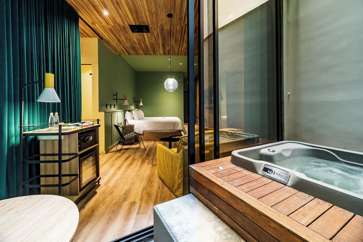 A modern hotel room with a wooden ceiling, a large window, a bed, a desk with a lamp, and a hot tub.
