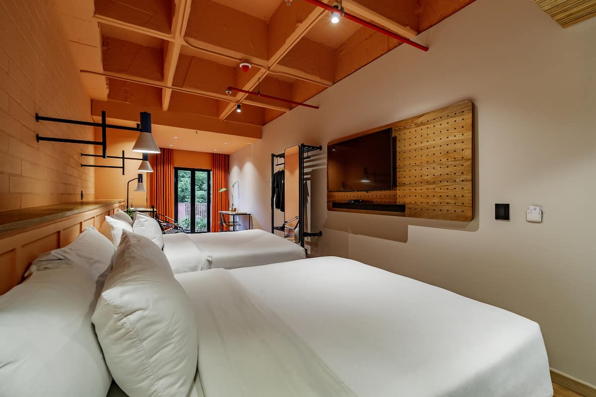 A hotel room with two beds, a TV mounted on the wall, a wooden ceiling, and a view of the outdoors through a window.