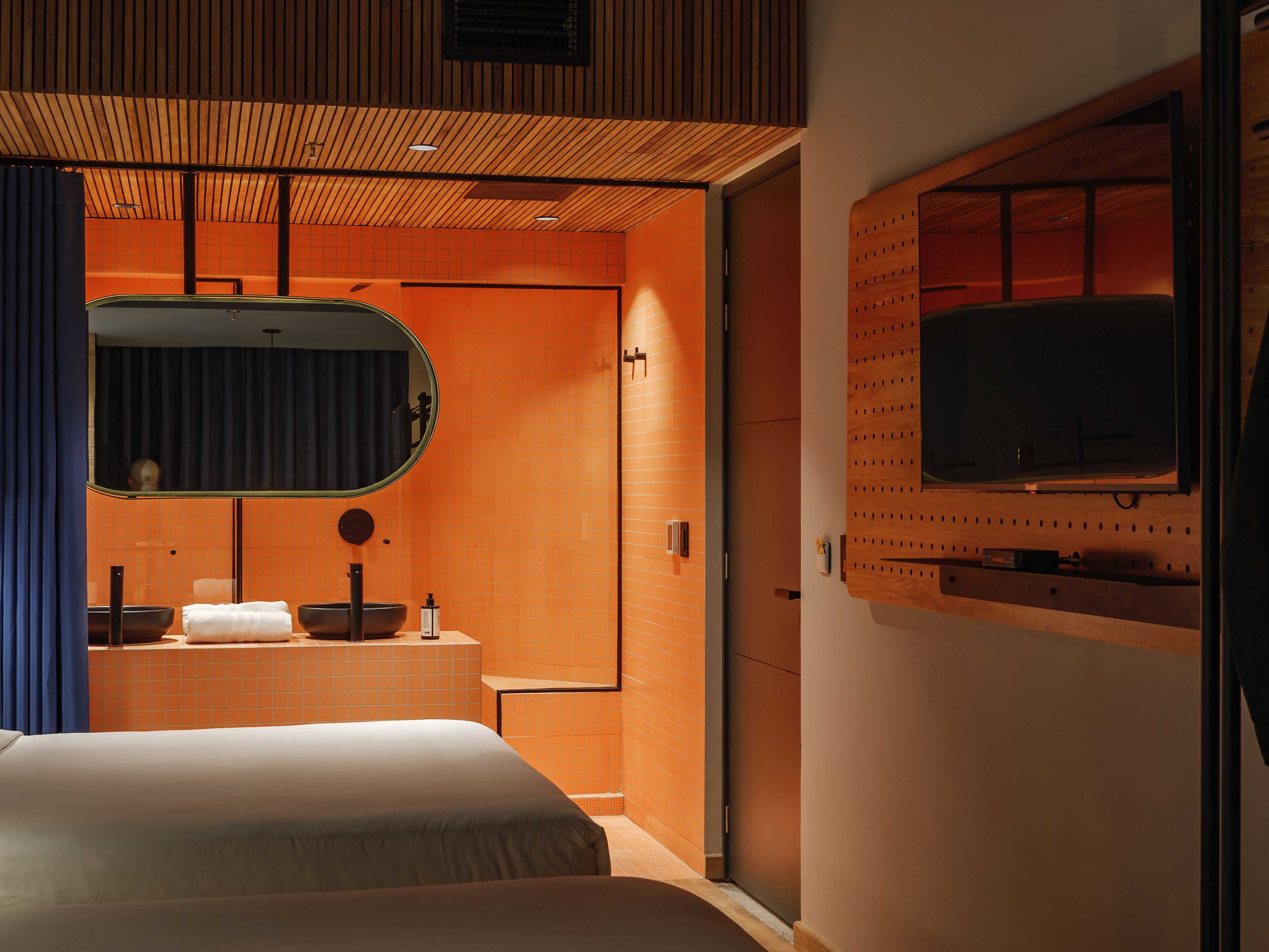 A modern hotel room with a large bed, a vanity with a mirror, a television mounted on the wall, and a wooden ceiling.