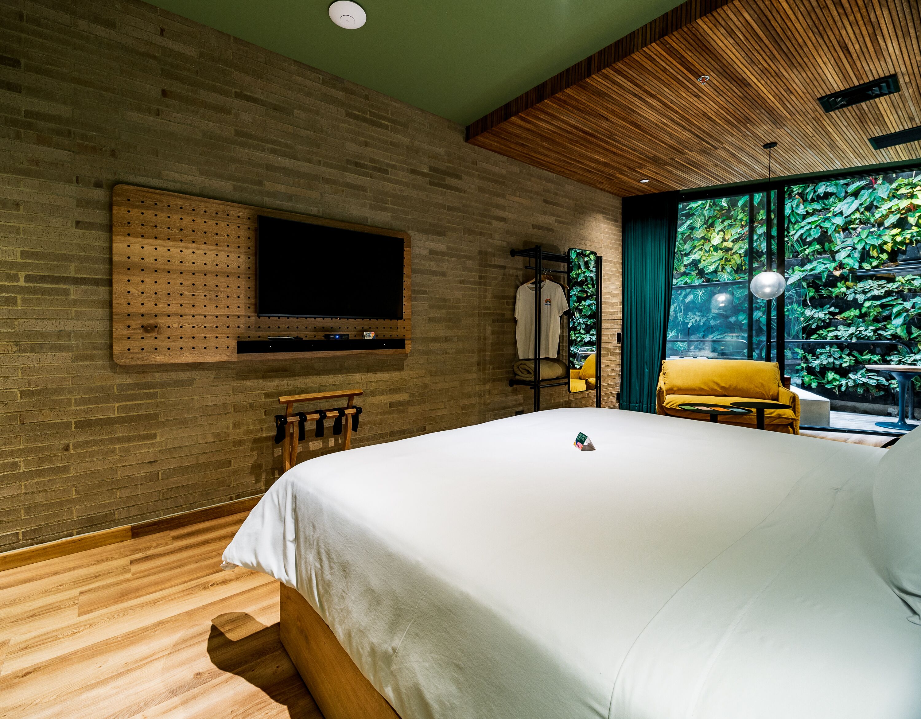 A modern hotel room with a large bed, wooden flooring, a mounted TV, and a view of greenery through large windows.