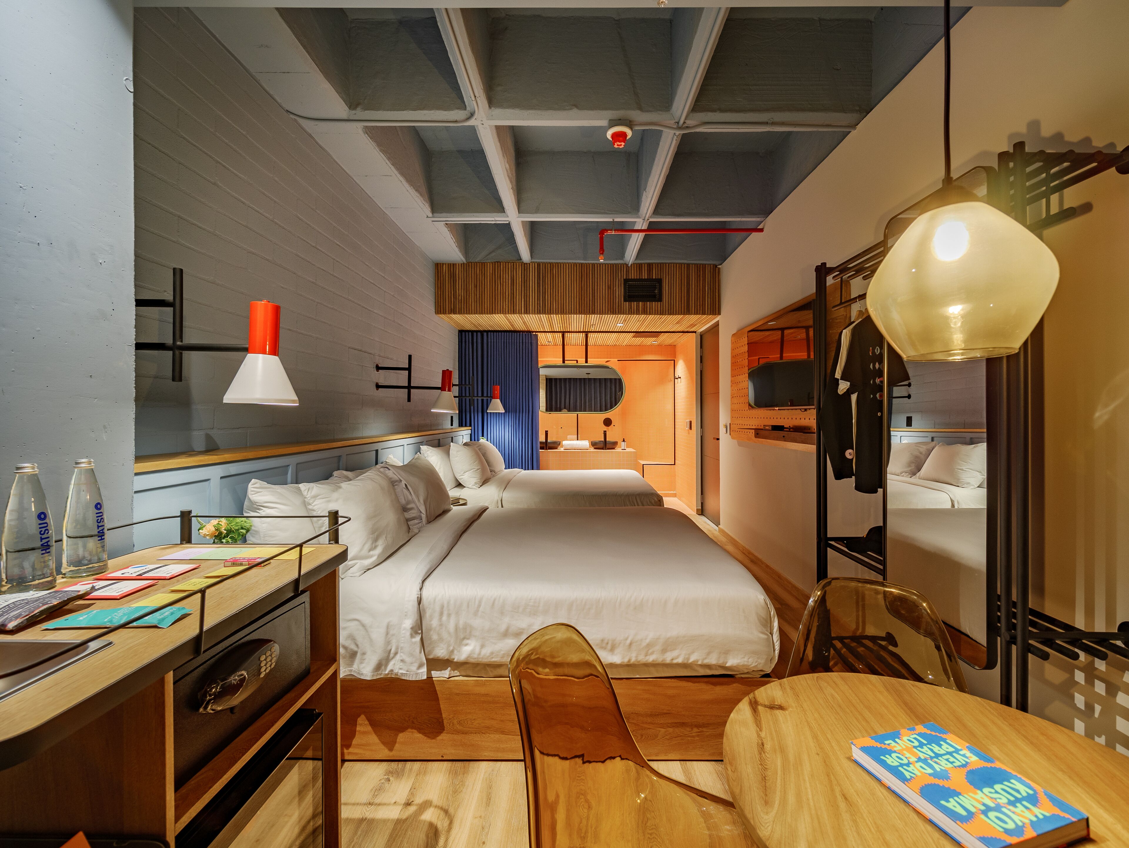 A hotel room with a bed, desk, chair, and a mirror. There is a window, a ceiling with exposed beams, and a hanging light fixture.