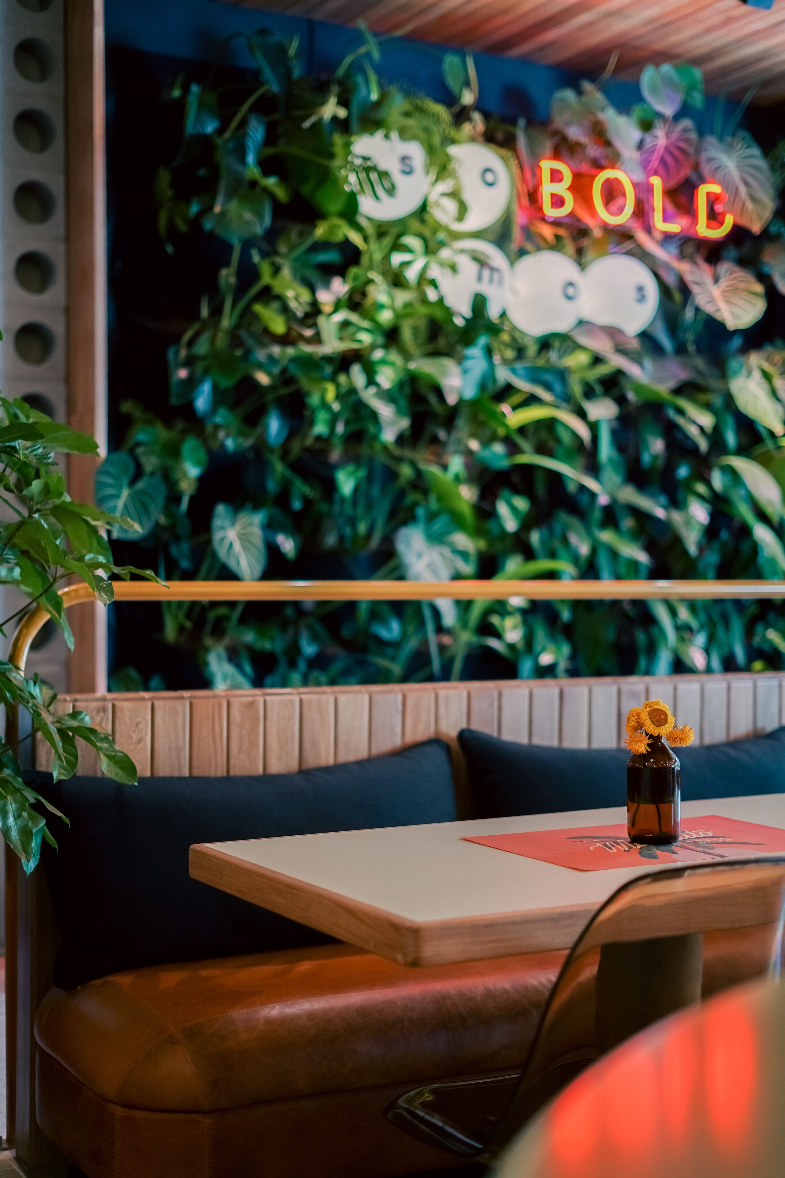 Interior de restaurante con mesa de madera, asientos tipo banqueta de cuero y una pared decorada con plantas verdes y un letrero de neón.
