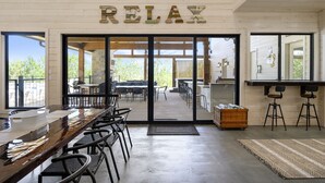 Ein geräumiger Innenbereich mit einem langen Holztisch und Stühlen, einer Küchenzeile mit Barhocker und einer großen Glasdoor, die zu einer Außenpatio führt.
