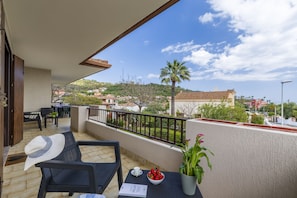 Un balcón con mobiliario exterior, una mesa con un frutero, y vistas a un pueblo con edificios y una palmera.
