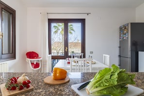 Una cocina con encimera de granito, una trona roja, una mesa de comedor con sillas y vistas a un espacio exterior con árboles a través de una puerta.
