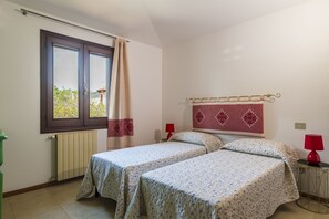 Une chambre avec deux lits, une fenêtre avec un rideau, un radiateur et deux lampes rouges posées sur des tables de chevet.