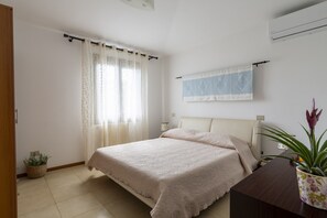 Une chambre à coucher comprenant un lit, une table de chevet, une plante en pot, une fenêtre avec des rideaux et un climatiseur fixé au mur.