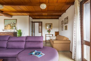 Ein Wohnzimmer mit einem violetten Sofa, einem braunen Sessel, Holzbalken an der Decke und einem Fenster mit Vorhängen.