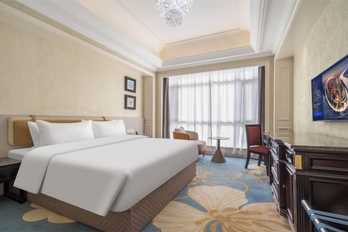 A hotel room with a large bed, a desk, a chair, a small table, and a large window with curtains.