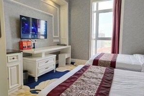 Habitación de hotel con una televisión de pantalla plana grande montada en la pared, un escritorio con cajones, dos camas con cubrecamas estampados y un ventanal con cortinas.