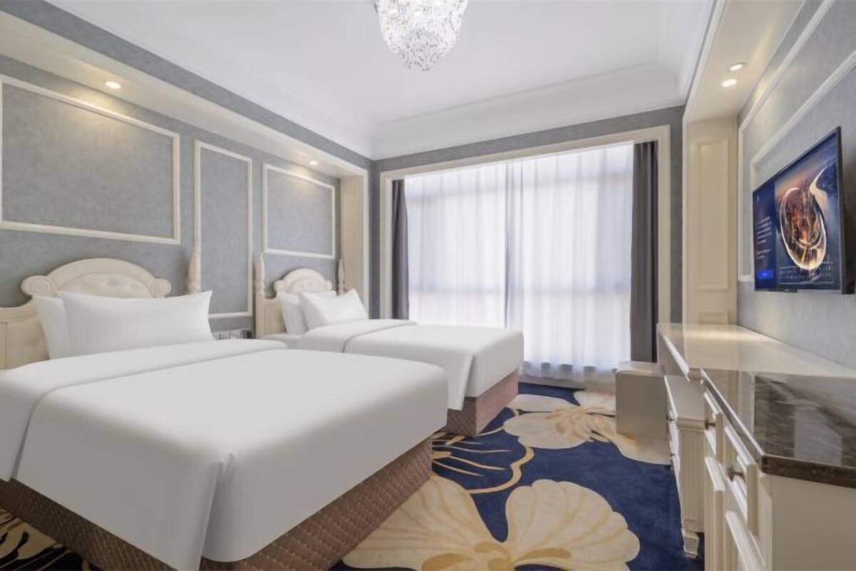 A hotel room with two beds, a large window with curtains, a flat-screen TV, and a decorative wall piece.