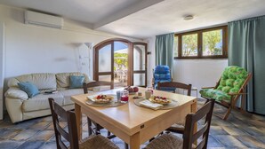 A dining area with a wooden table, chairs, and a sofa. There is a window with curtains, an air conditioning unit, and a view of the outdoors. - Tradewinds Family House (Tortolì)