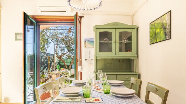 A dining area with a table set for four, a green cabinet, and a view through an open door to the outside. - Vacation House Summer By The Sea (Tortoli')