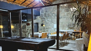 A pool table in a room with stone walls and a wooden ceiling. There are tables and chairs outside the room, and a plant is visible. - Alqueiturismo - Garden House (Corujeira)