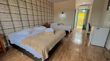 A hotel room with two beds, a tiled floor, a wall with a textured pattern, a small table with a lamp, and a refrigerator.