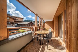 Un balcon en bois avec une table ronde et des chaises, d’où l’on peut admirer un village aux bâtiments traditionnels et, au loin, des montagnes.